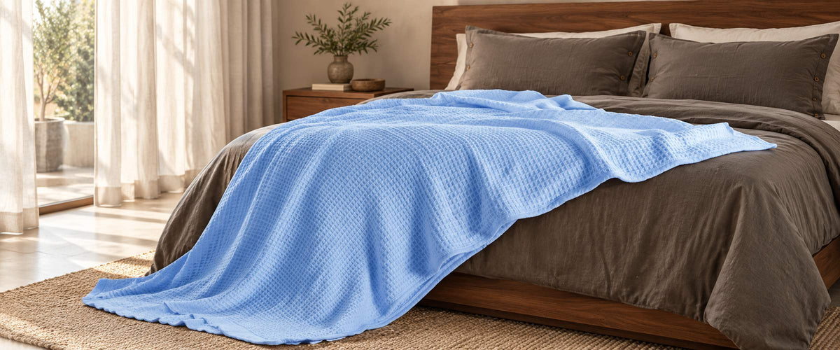 Light blue waffle-weave cotton blanket draped across taupe bedding in a sunlit bedroom