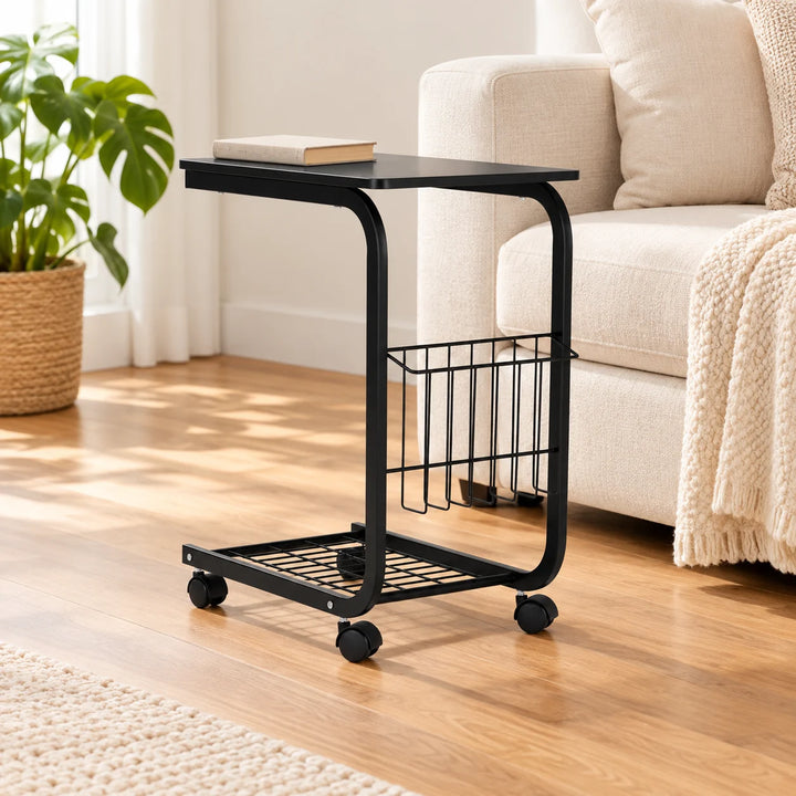 Black rolling laptop side table with magazine basket and mesh base beside a cream sofa and natural plant