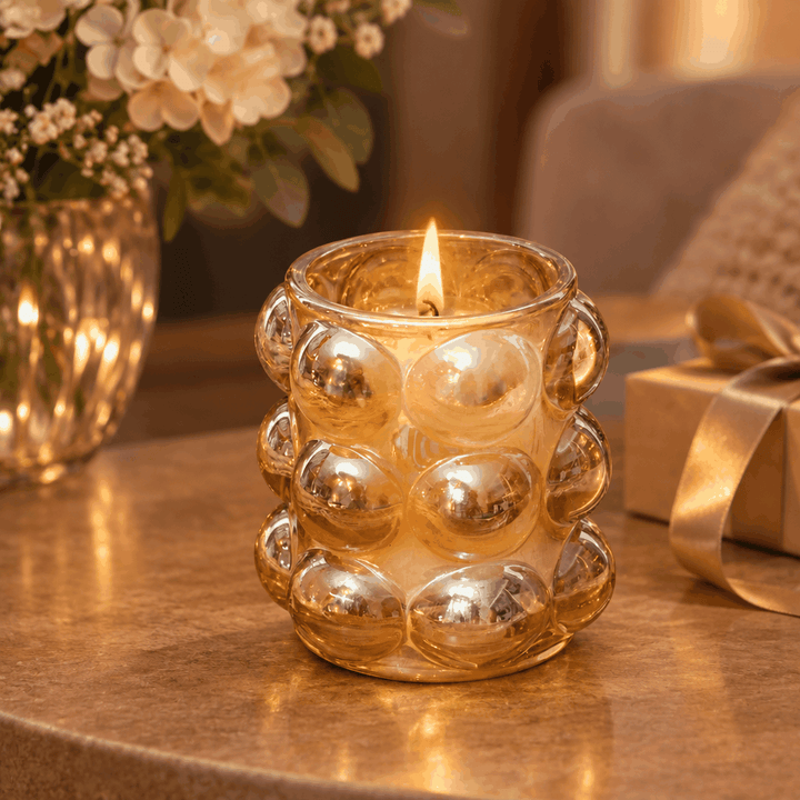 Lit candle in amber bubble glass holder on table with flowers and gift box