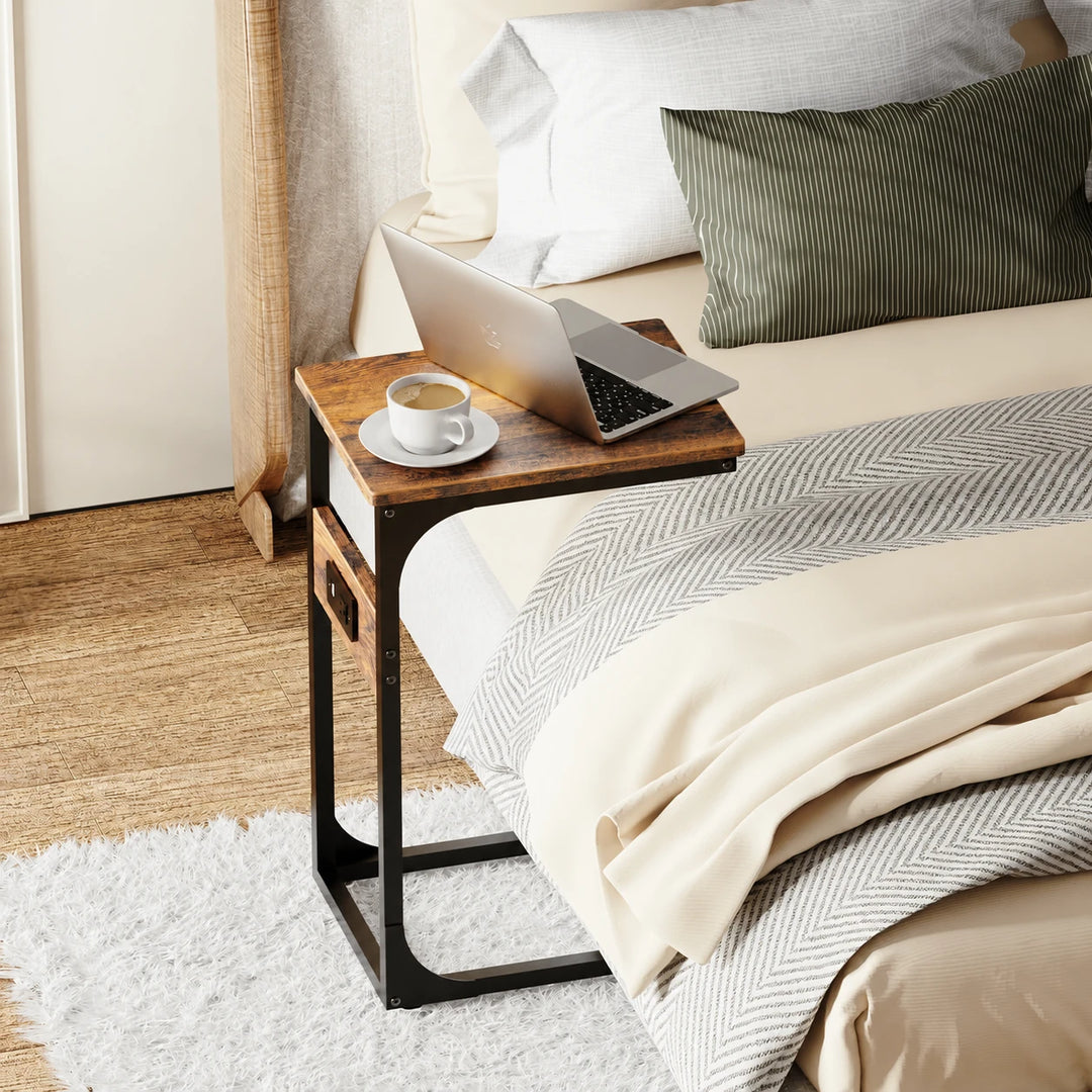 One rustic brown C-shaped side table with black frame and power socket, holding a laptop and cup beside a bed