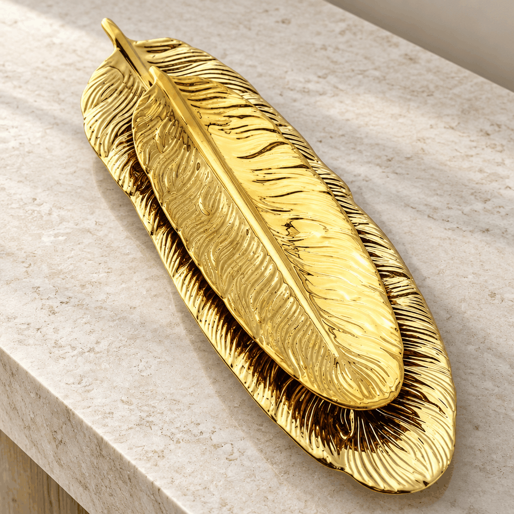 Two golden feather jewellery plates stacked on marble surface, close-up angled view