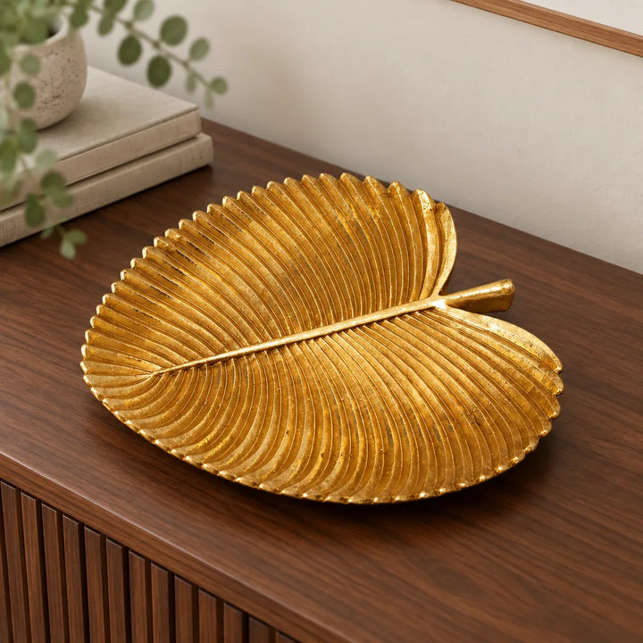Wide golden leaf shaped tray on dark wooden sideboard with plant behind