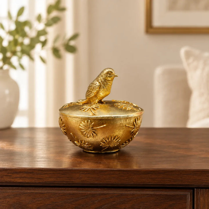 Golden storage bowl with bird figurine on lid placed on wooden sideboard