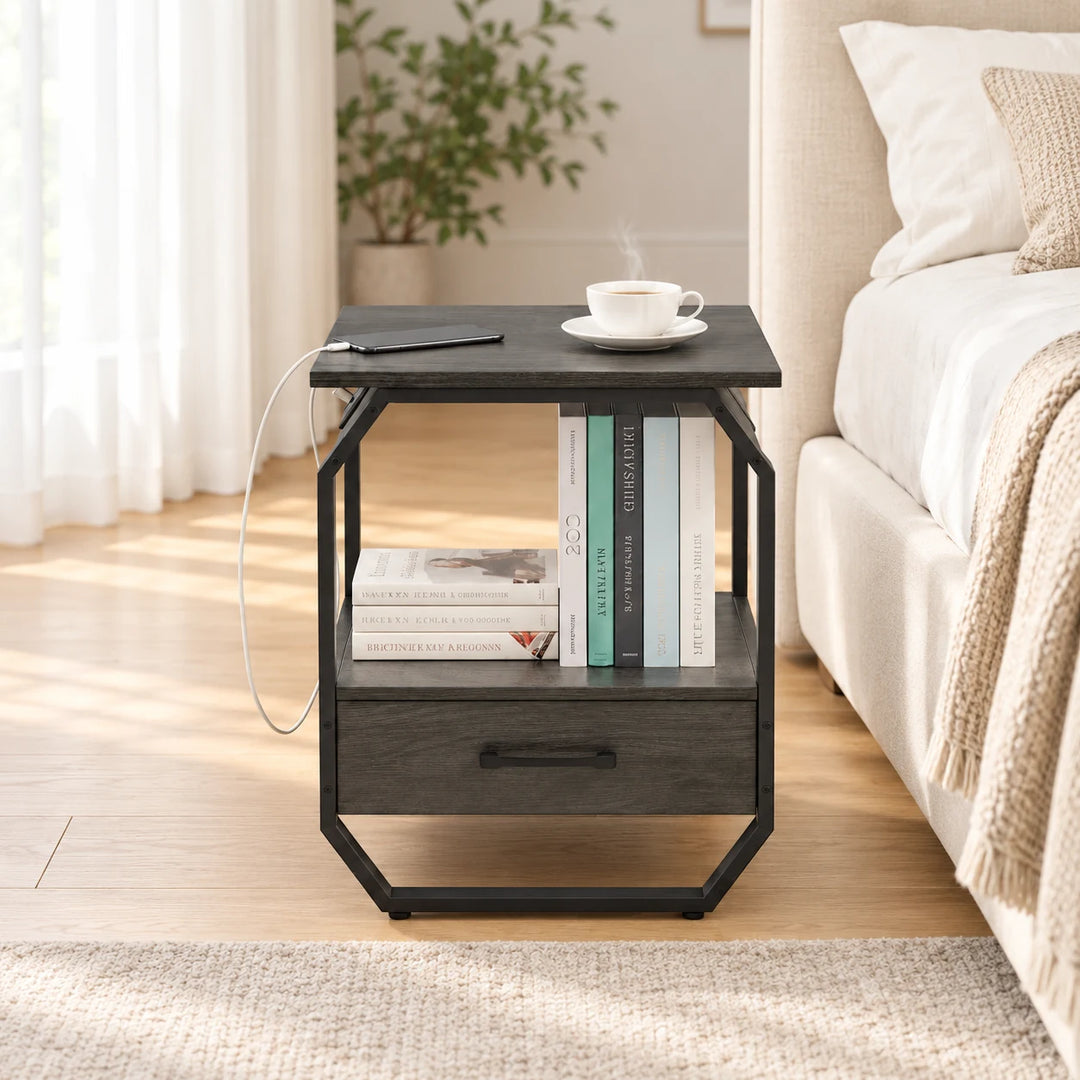 Hexi charcoal bedside table with angular black frame, open shelf and drawer beside a cream bed in natural daylight