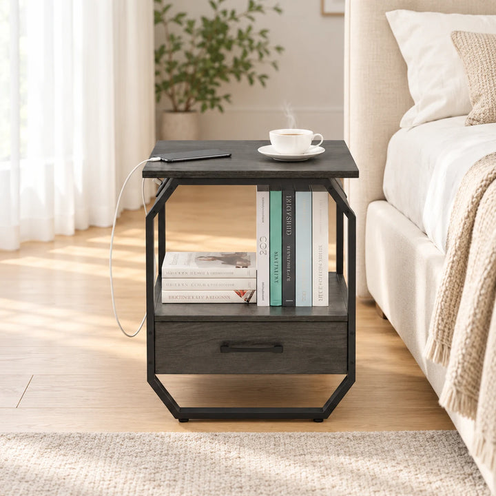 Hexi charcoal bedside table with angular black frame, open shelf and drawer beside a cream bed in natural daylight