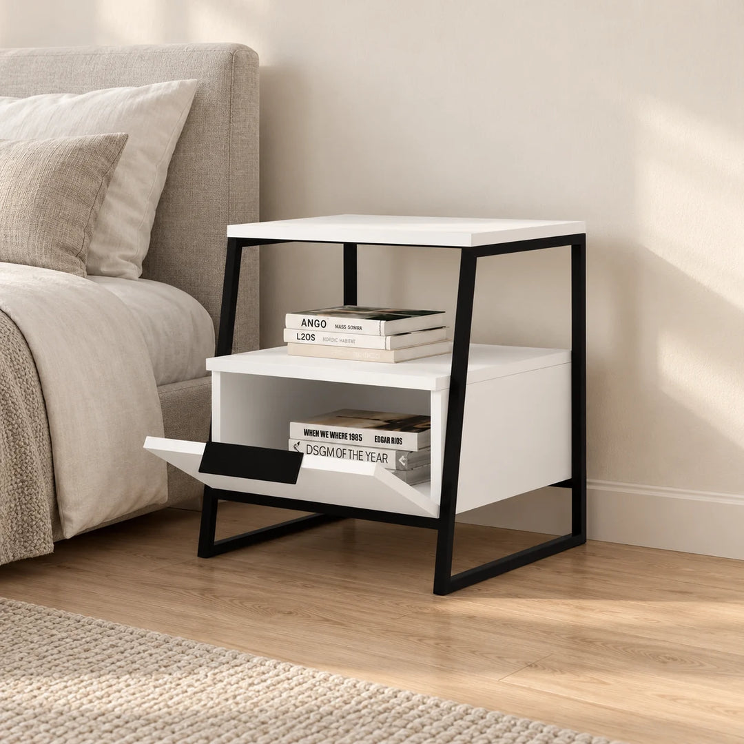 White Hokku bedside table with black angled metal frame, open shelf and drop-down storage flap beside a neutral upholstered bed.