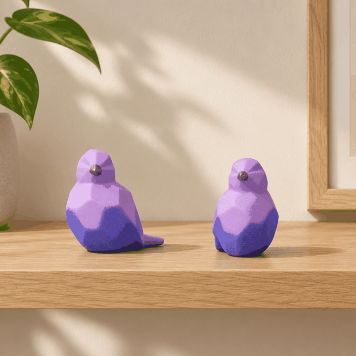 Pair of purple geometric bird decor figurines on wooden shelf with leafy plant