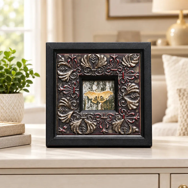 Small square black photo frame with embossed floral border beside plant