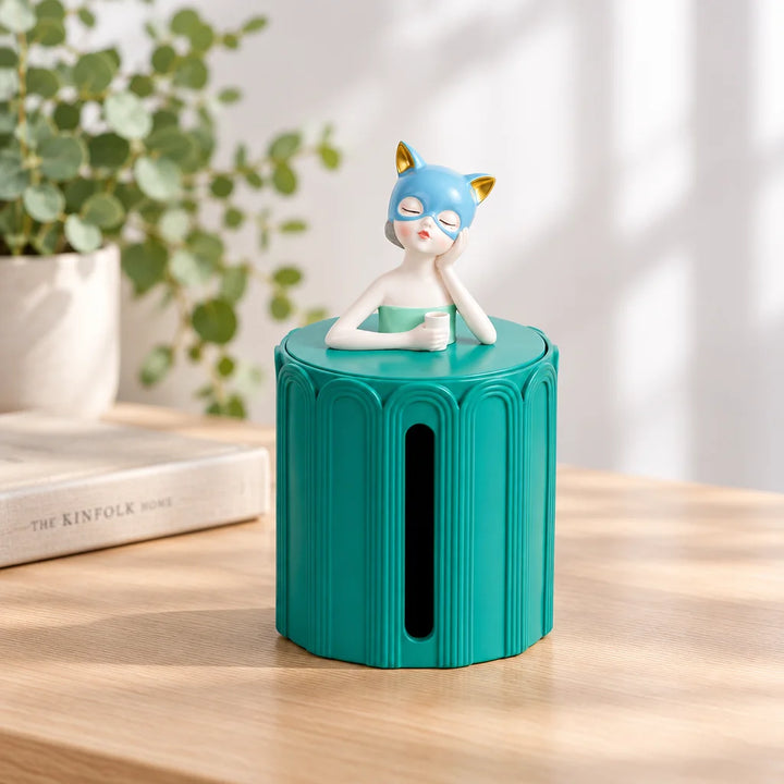 Turquoise round tissue box with girl figurine on lid on wooden desk beside plant