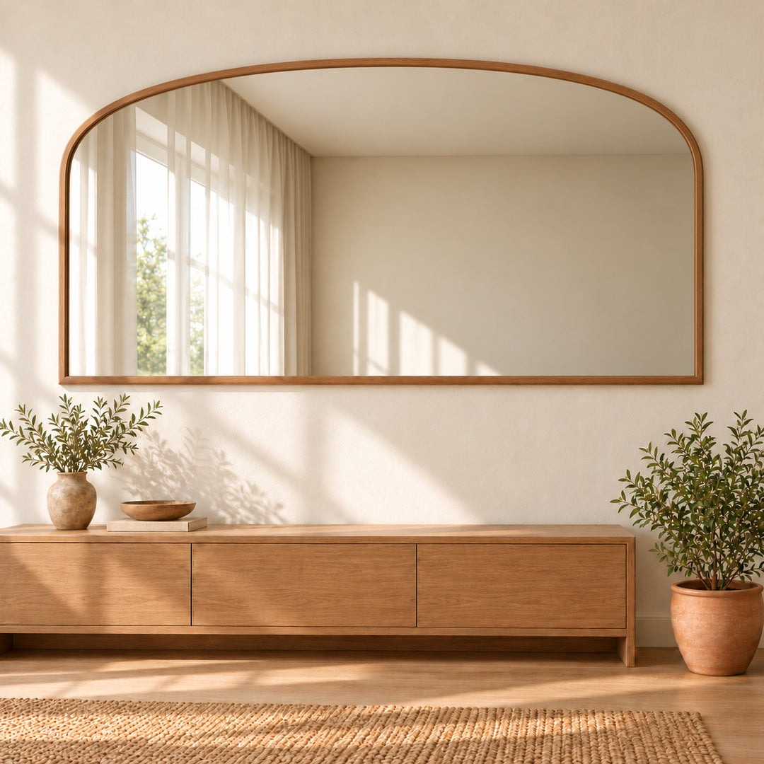 Wide horizontal arched mirror with wooden frame above a sideboard beside greenery.