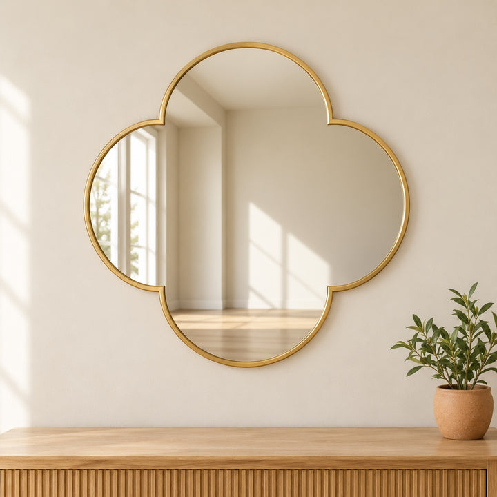 Square-proportioned four-lobed gold tone quatrefoil mirror in a digitally styled room with greenery.