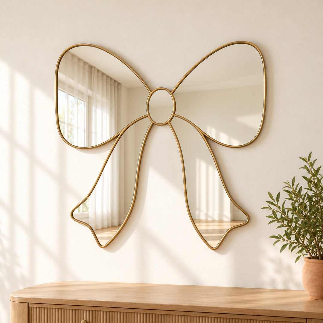 Bow ribbon wall mirror with gold tone loops and tails beside potted greenery.