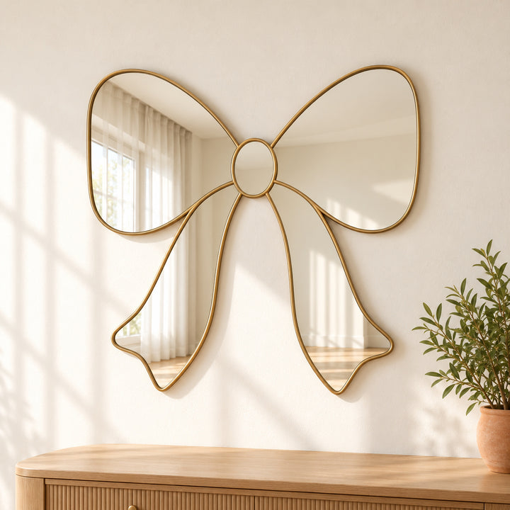 Bow ribbon wall mirror with gold tone loops and tails beside potted greenery.