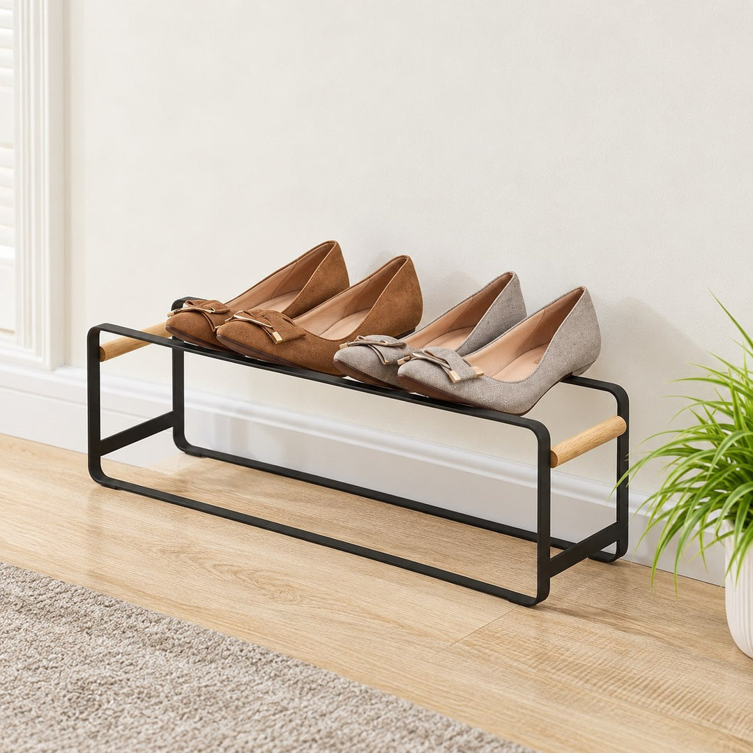 One Kiru black metal shoe rack unit with natural wood side handles and two pairs of shoes against a wall